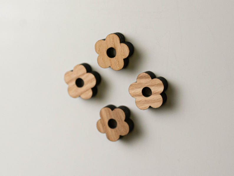 Puede incluir: Cuatro imanes de madera con forma de flor. Cada im&aacute;n tiene un dise&ntilde;o de flor marr&oacute;n claro con un borde marr&oacute;n oscuro y un agujero circular en el centro. Los imanes est&aacute;n dispuestos sobre una superficie blanca.