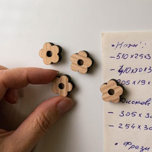 Puede incluir: Cuatro imanes de madera con forma de flor, con centro marr&oacute;n claro y contorno marr&oacute;n oscuro. Una mano sostiene un im&aacute;n. Una nota manuscrita con medidas est&aacute; en el fondo.