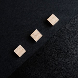 May include: Three small wooden squares arranged in a row on a black surface. The squares are light brown and have a smooth finish.