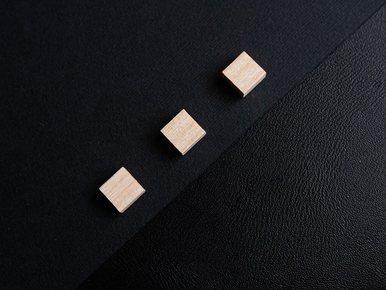 May include: Three small wooden squares arranged in a row on a black surface. The squares are light brown and have a smooth finish.