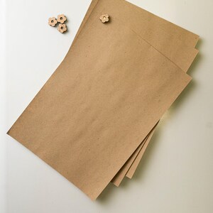 Puede incluir: Una pila de tres hojas rectangulares de papel kraft marr&oacute;n con un chincheta de madera en forma de flor. Tres adornos de madera adicionales en forma de flor est&aacute;n dispersos cerca.