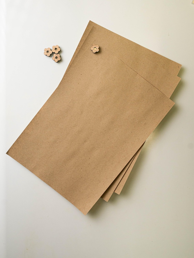 Puede incluir: Una pila de tres hojas rectangulares de papel kraft marr&oacute;n con un chincheta de madera en forma de flor. Tres adornos de madera adicionales en forma de flor est&aacute;n dispersos cerca.