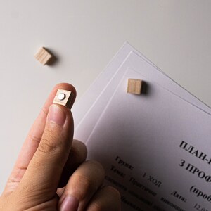 May include: A close-up of a hand holding a small wooden cube with a silver circle on the top. The cube is being held over a white surface with a stack of papers on it. The papers have text on them that reads "PLAN-KONSPE Z PROFMAYSTR (probne zavd) 1 ход Група: Тема: Практичне виконання елемента консдкові ди Дата: 12.02"