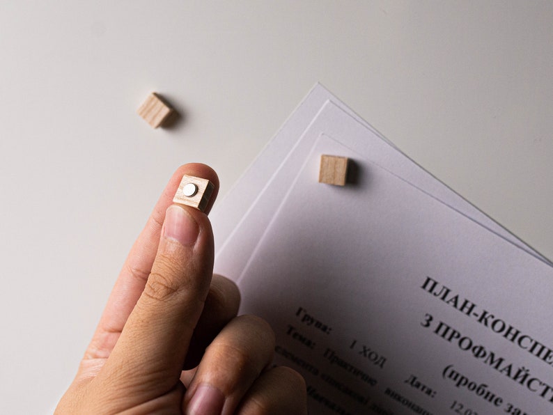 May include: A close-up of a hand holding a small wooden cube with a silver circle on the top. The cube is being held over a white surface with a stack of papers on it. The papers have text on them that reads "PLAN-KONSPE Z PROFMAYSTR (probne zavd) 1 ход Група: Тема: Практичне виконання елемента консдкові ди Дата: 12.02"