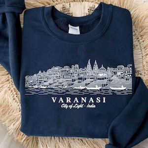 Pode incluir: Uma camisola azul-marinho de gola redonda com uma ilustração de linhas brancas do horizonte da cidade de Varanasi ao longo do rio Ganges com barcos. Abaixo da ilustração, o texto diz VARANASI City of Light - India em letras brancas.