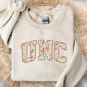 K&ouml;nnte beinhalten: Cremefarbenes Sweatshirt mit den Buchstaben "UNC" in einem floralen Design. Die Buchstaben sind braun umrandet und mit rosa, pfirsichfarbenen und gr&uuml;nen Blumenmustern gef&uuml;llt. Das Sweatshirt liegt auf einer gewebten, beigefarbenen Oberfl&auml;che.