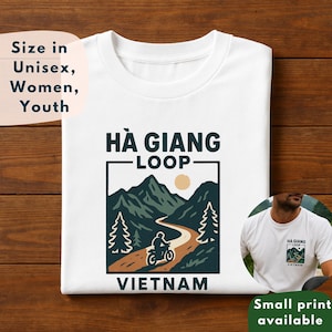 Puede incluir: Camiseta blanca con un gráfico de una motocicleta en una carretera sinuosa a través de montañas, con el texto "HÀ GIANG LOOP VIETNAM". Disponible en tallas unisex, de mujer y juvenil.