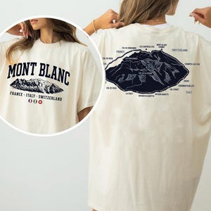 May include: An off-white t-shirt featuring a dark blue graphic of the Mont Blanc mountain range. The front displays the text MONT BLANC, FRANCE - ITALY - SWITZERLAND, and three small flags. The back shows a detailed map of the mountain peaks.