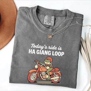Puede incluir: Una camiseta gris con el texto "Today's ride is HA GIANG LOOP" y una rana de dibujos animados montada en una moto naranja. La camiseta tiene cuello redondo y una pequeña etiqueta. Un collar plateado con un colgante está sobre la camiseta.