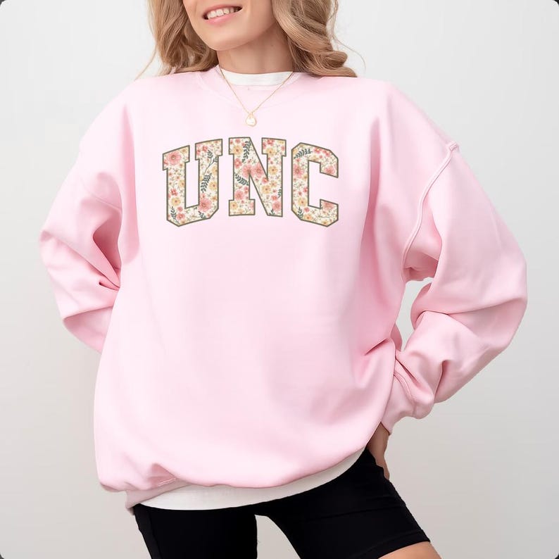 K&ouml;nnte beinhalten: Ein hellrosa Sweatshirt mit den Buchstaben "UNC" auf der Brust. Die Buchstaben sind mit einem Blumenmuster in Rosa-, Gelb- und Gr&uuml;nt&ouml;nen gef&uuml;llt. Das Sweatshirt hat einen Rundhalsausschnitt.