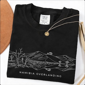 Peut inclure: T-shirt noir avec un motif blanc d'un paysage désertique et l'inscription "NAMIBIA OVERLANDING". Un collier doré avec un pendentif rond est posé sur le t-shirt. Le t-shirt a une étiquette Comfort Colors.