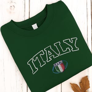 Könnte beinhalten: Ein dunkelgrünes Sweatshirt mit dem Wort "ITALY" in weißer, bogenförmiger Schrift gestickt. Darunter befindet sich ein kleines Emblem mit den Farben der italienischen Flagge. Das Sweatshirt liegt gefaltet auf einer weißen Holzoberfläche.