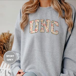 K&ouml;nnte beinhalten: Ein hellgraues Sweatshirt mit den Buchstaben "UNC" in einem floralen Design. Das Sweatshirt hat einen Rundhalsausschnitt und lange &Auml;rmel. Der Text "SPORT GREY" befindet sich in einem Kreis auf der linken Seite. Ein l&auml;ssiges Sweatshirt.