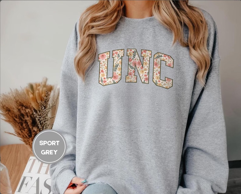 K&ouml;nnte beinhalten: Ein hellgraues Sweatshirt mit den Buchstaben "UNC" in einem floralen Design. Das Sweatshirt hat einen Rundhalsausschnitt und lange &Auml;rmel. Der Text "SPORT GREY" befindet sich in einem Kreis auf der linken Seite. Ein l&auml;ssiges Sweatshirt.
