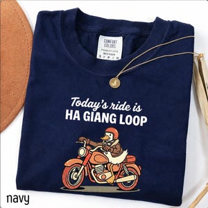 Może przedstawiać: Granatowa koszulka z napisem "Today's ride is HA GIANG LOOP" i rysunkiem kaczki na motocyklu. Złoty naszyjnik z wisiorkiem leży na koszulce. Koszulka ma metkę Comfort Colors.