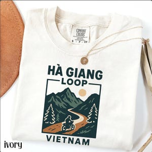 May include: Ivory-colored t-shirt with a graphic of a motorcycle on a winding road through mountains, with the text "Hà Giang Loop Vietnam." The shirt has a crew neck and short sleeves. A gold necklace with a pendant is draped across the shirt.