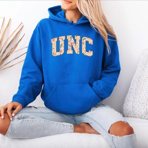 K&ouml;nnte beinhalten: Ein k&ouml;nigsblauer Kapuzenpullover mit den Buchstaben "UNC" in einem floralen Muster auf der Brust. Der Pullover hat eine Vordertasche und eine Kapuze mit Kordelzug. Das Model tr&auml;gt hellblaue Jeans.
