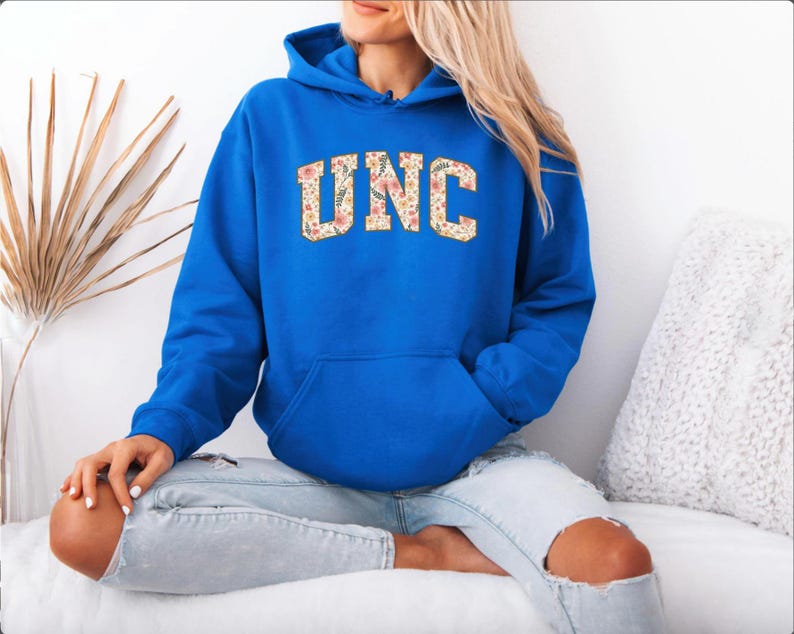 K&ouml;nnte beinhalten: Ein k&ouml;nigsblauer Kapuzenpullover mit den Buchstaben "UNC" in einem floralen Muster auf der Brust. Der Pullover hat eine Vordertasche und eine Kapuze mit Kordelzug. Das Model tr&auml;gt hellblaue Jeans.