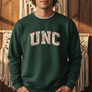 K&ouml;nnte beinhalten: Ein dunkelgr&uuml;nes Sweatshirt mit den Buchstaben "UNC" in einem floralen Design auf der Brust. Das Sweatshirt hat einen Rundhalsausschnitt und lange &Auml;rmel. Der Hintergrund ist eine Holzwand mit dekorativen Elementen.