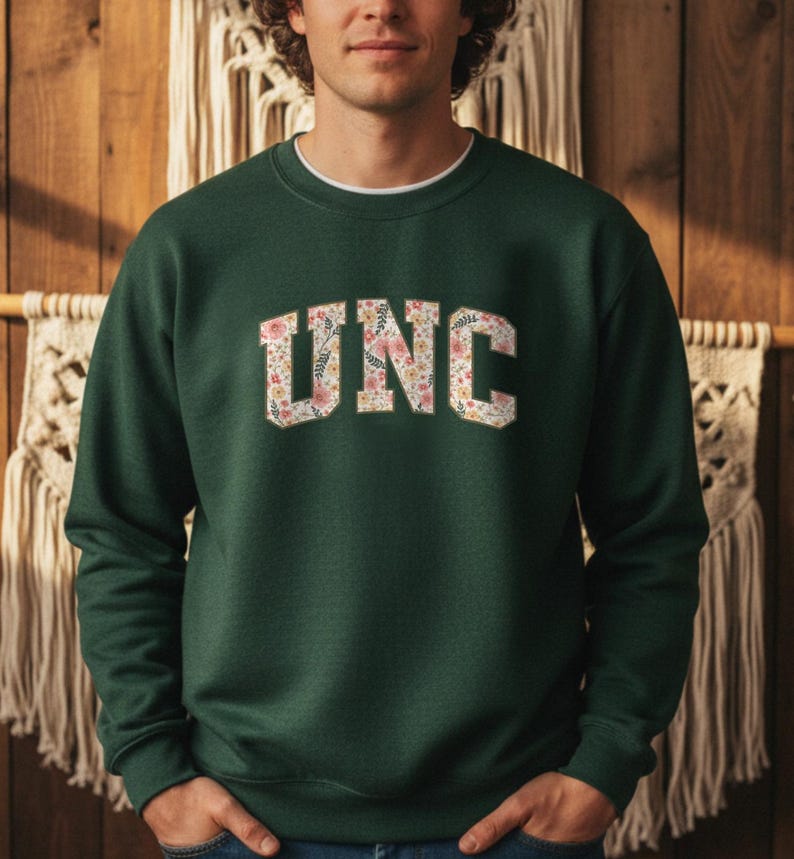 K&ouml;nnte beinhalten: Ein dunkelgr&uuml;nes Sweatshirt mit den Buchstaben "UNC" in einem floralen Design auf der Brust. Das Sweatshirt hat einen Rundhalsausschnitt und lange &Auml;rmel. Der Hintergrund ist eine Holzwand mit dekorativen Elementen.