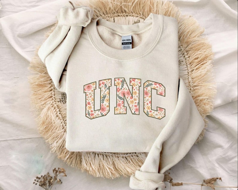 K&ouml;nnte beinhalten: Cremefarbenes Sweatshirt mit den Buchstaben "UNC" in einem floralen Design. Die Buchstaben sind braun umrandet und mit rosa, pfirsichfarbenen und gr&uuml;nen Blumenmustern gef&uuml;llt. Das Sweatshirt liegt auf einer gewebten, beigefarbenen Oberfl&auml;che.