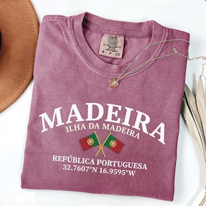 May include: A mauve Comfort Colors t-shirt with the word "MADEIRA" in white block letters. Below the word "MADEIRA" is "ILHA DA MADEIRA" and "REPÚBLICA PORTUGUESA". Two crossed flags and coordinates are also printed on the shirt. A gold necklace is draped across the shirt.
