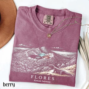 May include: A berry-colored short-sleeve t-shirt featuring a graphic print of a Komodo dragon in a volcanic landscape. The text "FLORES Komodo Indonesia" is printed below the graphic. The shirt has a Comfort Colors brand label at the neck.