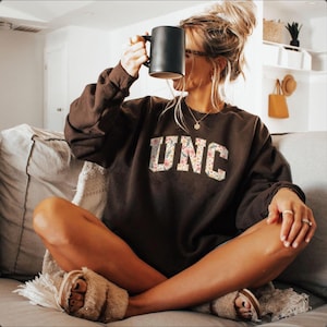 K&ouml;nnte beinhalten: Ein braunes Sweatshirt mit den Buchstaben "UNC" in einem floralen Design. Die Person h&auml;lt eine schwarze Tasse. Die Person tr&auml;gt flauschige Hausschuhe und sitzt auf einem Sofa.