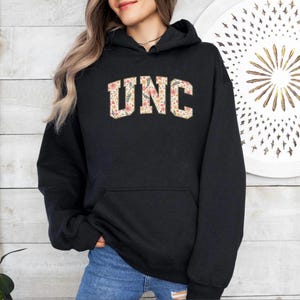 K&ouml;nnte beinhalten: Schwarzer Kapuzenpullover mit den Buchstaben "UNC" in einem floralen Muster auf der Brust. Der Pullover hat eine Vordertasche und eine Kapuze mit Kordelzug. Die Buchstaben sind in einer Schriftart im College-Stil.