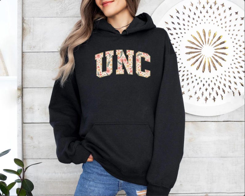 K&ouml;nnte beinhalten: Schwarzer Kapuzenpullover mit den Buchstaben "UNC" in einem floralen Muster auf der Brust. Der Pullover hat eine Vordertasche und eine Kapuze mit Kordelzug. Die Buchstaben sind in einer Schriftart im College-Stil.