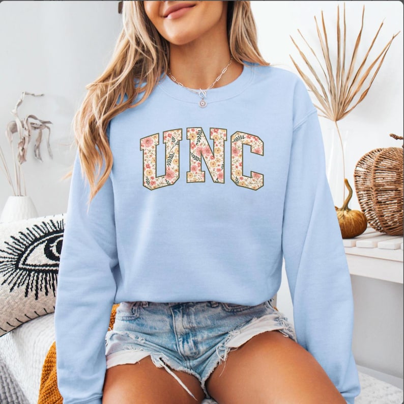K&ouml;nnte beinhalten: Hellblaues Sweatshirt mit den Buchstaben "UNC" in einem floralen Design. Das Sweatshirt hat lange &Auml;rmel und einen Rundhalsausschnitt. Das florale Design umfasst rosa, gelbe und wei&szlig;e Blumen. Das Sweatshirt ist aus einem weichen Material.