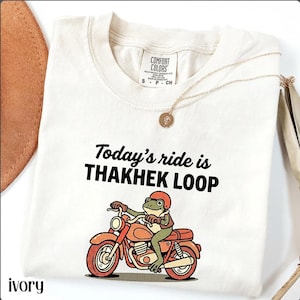 Może przedstawiać: Kremowa koszulka z napisem "Today's ride is THAKHEK LOOP" i kreskówkową żabą na pomarańczowym motocyklu. Żaba ma na sobie kask. Złoty naszyjnik jest zarzucony na koszulkę.