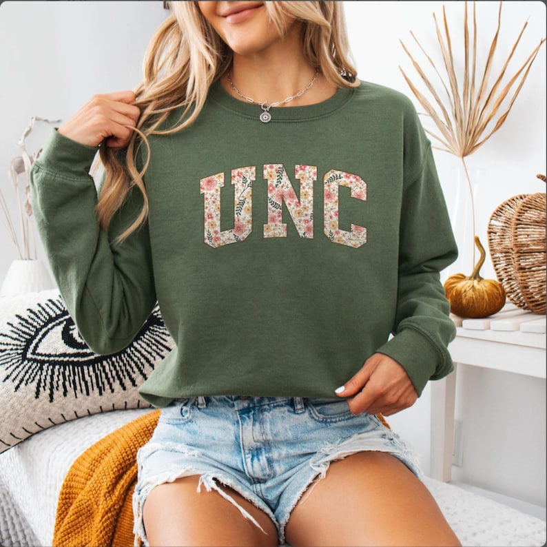 K&ouml;nnte beinhalten: Olivgr&uuml;nes Sweatshirt mit den Buchstaben "UNC" in einem floralen Muster. Das lang&auml;rmlige Oberteil ist aus einem weichen Material gefertigt. Das Sweatshirt ist ein l&auml;ssiger Stil.