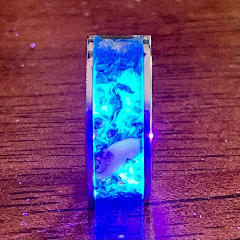 Pu&ograve; includere: Un anello rettangolare color argento con un centro in resina blu brillante. La resina contiene piccoli elementi decorativi blu e bianchi e un oggetto bianco a forma di conchiglia. L'anello &egrave; illuminato da una luce blu.