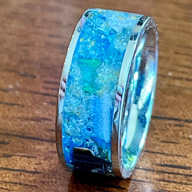 Pu&ograve; includere: Un anello color argento con una fascia larga. La fascia &egrave; riempita con un mix di resina blu, turchese e bianca, creando un aspetto strutturato simile all'oceano. L'anello &egrave; appoggiato su una superficie di legno.