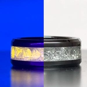 May include: A black ring with a band of yellow and silver-colored fragments. The ring has a polished, reflective surface. The fragments are encased in a clear material, creating a unique design.