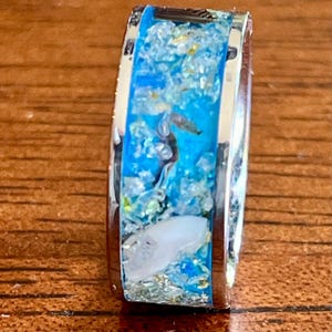 Pu&ograve; includere: Un anello d'argento con un centro in resina blu, con un cavalluccio marino in miniatura e altri elementi a tema marino. La fascia dell'anello &egrave; in argento lucido e la resina &egrave; riempita di glitter e piccoli elementi decorativi. Un gioiello unico.