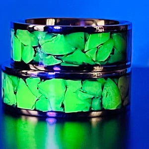 May include: Two stacked metal rings featuring a central inlay of glowing green, jagged stone fragments. The metallic bands have a polished finish that reflects light, while the embedded stone pieces emit a bright, luminous green light.