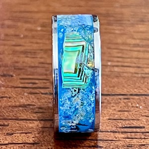 Pu&ograve; includere: Primo piano di un anello d'argento con un'intarsio blu e iridescente vibrante. L'intarsio presenta un mix di colori blu, verdi e bianchi, creando un design unico e accattivante. L'anello &egrave; posto su uno sfondo di legno.