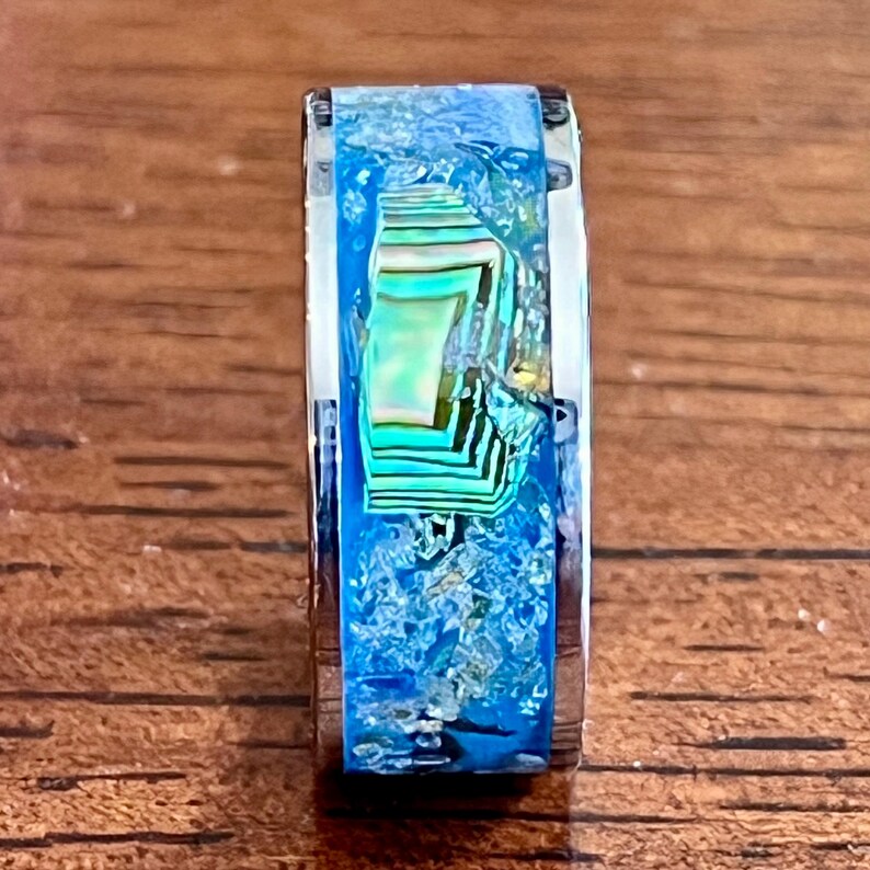 Pu&ograve; includere: Primo piano di un anello d'argento con un'intarsio blu e iridescente vibrante. L'intarsio presenta un mix di colori blu, verdi e bianchi, creando un design unico e accattivante. L'anello &egrave; posto su uno sfondo di legno.