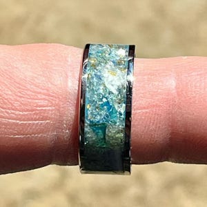 Pu&ograve; includere: Primo piano di un anello con fascia nera e intarsio colorato. L'intarsio presenta sfumature di blu, verde e oro, creando un aspetto strutturato simile all'oceano. L'anello &egrave; indossato su un dito.