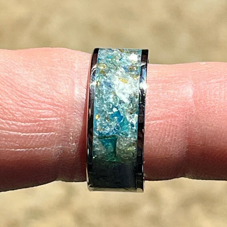 Pu&ograve; includere: Primo piano di un anello con fascia nera e intarsio colorato. L'intarsio presenta sfumature di blu, verde e oro, creando un aspetto strutturato simile all'oceano. L'anello &egrave; indossato su un dito.