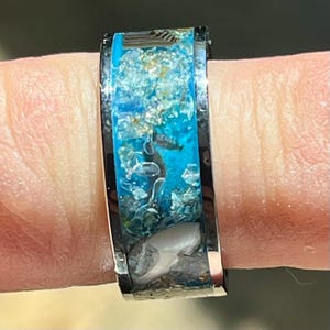 Pu&ograve; includere: Un anello nero con un design rettangolare. Il centro dell'anello presenta una vibrante resina turchese riempita di piccoli frammenti chiari e argentati, insieme a un piccolo oggetto bianco a forma di dente. L'anello &egrave; tenuto da un dito.