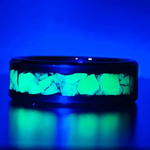 May include: A black ring with a band of glowing green stones. The ring is set against a dark blue background, with the green stones reflecting on the surface below. The ring's design is modern and eye-catching.
