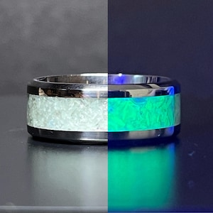 May include: A silver-toned ring with a wide band. The ring's center section is filled with a light-colored, textured material that glows bright green on one side. The other side of the ring is not illuminated.