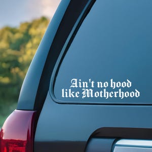 May include: White vinyl decal with the text "Ain't no hood like Motherhood" in a gothic font, applied to the back window of a dark blue car.