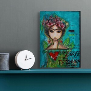 May include: An artistic painting of a woman with a floral crown, a red heart, and text in Spanish. The artwork is on a teal background. A white clock and a gray container are on a shelf.