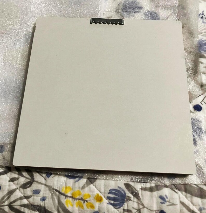 May include: A rectangular, off-white canvas with a metal sawtooth hanger attached to the top. The canvas is set against a patterned fabric background. The canvas is ready to be hung.