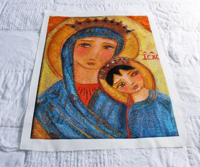 May include: A framed religious icon featuring a mother and child, rendered in a folk art style. The figures are depicted with soft features and are wearing blue and red garments. The background is gold with the Greek letters IC XC.