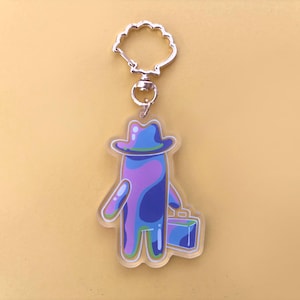 Puede incluir: Un llavero acrílico transparente con una figura de dibujos animados en azul y morado, con un sombrero azul y un maletín azul. El llavero tiene un cierre plateado.